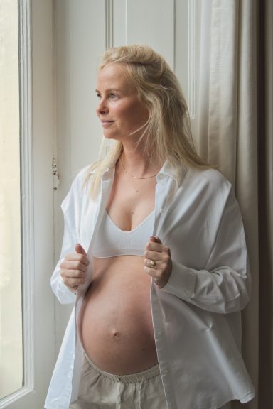 Zwangere vrouw in witte top, voor een raam, kijkt naar buiten.